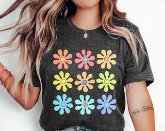 May include: A dark grey heather t-shirt with a colorful daisy print. The daisies are in a 3x3 grid pattern and are in shades of pink, blue, yellow, and orange. The text "More Ocean" is written on the right sleeve of the shirt.