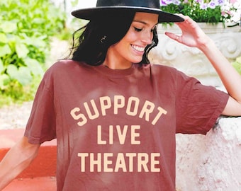 May include: A woman wearing a maroon t-shirt with the text "Support Live Theatre" in a light orange color. She is wearing a black hat and is sitting on a red brick surface.