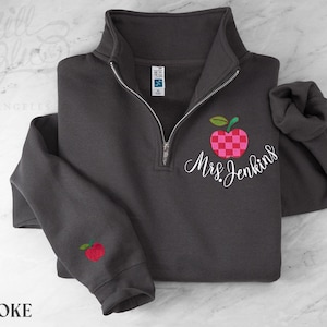 May include: A charcoal gray quarter-zip sweatshirt featuring an embroidered red and pink checkered apple design on the chest with the white cursive text Mrs. Jenkins below it and a smaller matching apple embroidered on the left sleeve cuff.