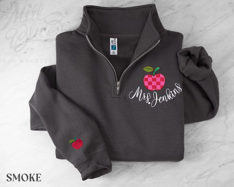 May include: A charcoal gray quarter-zip sweatshirt featuring an embroidered red and pink checkered apple design on the chest with the white cursive text Mrs. Jenkins below it and a smaller matching apple embroidered on the left sleeve cuff.
