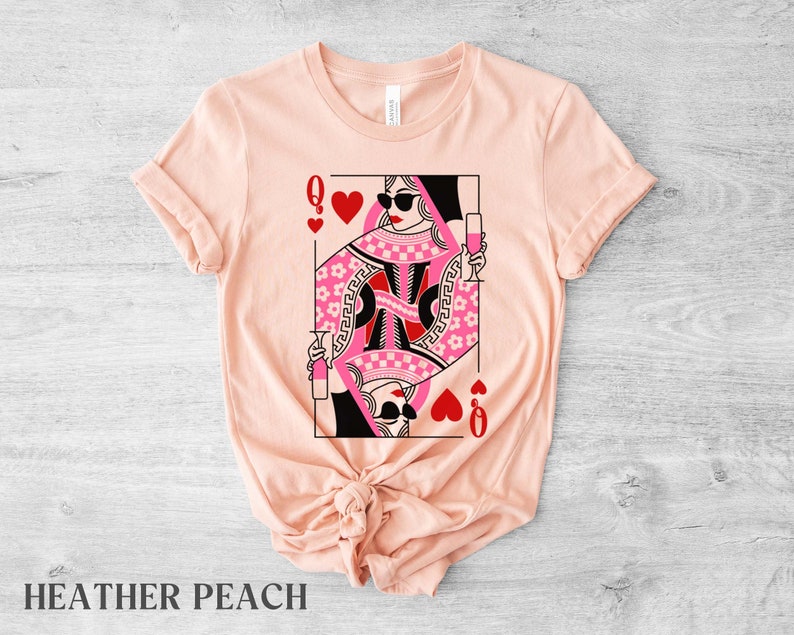 K&ouml;nnte beinhalten: Ein T-Shirt in Pfirsichfarbe mit einem Aufdruck der Herzdame aus einem Kartenspiel. Die Dame tr&auml;gt eine Sonnenbrille und h&auml;lt ein Glas Wein in der Hand. Der Text "Q" und ein Herz sind auf der Karte.