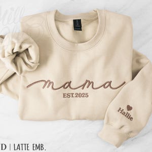 May include: A sand-colored crewneck sweatshirt featuring brown embroidery on the chest that reads "mama" in a cursive script with "EST. 2025" underneath. The left sleeve is embroidered with a small brown heart and the name "Hallie".