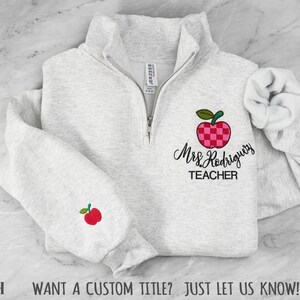 May include: A light gray heather quarter-zip sweatshirt featuring an embroidered red and pink checkered apple design on the chest with the text Mrs. Rodriguez TEACHER below it, and a small red apple embroidered on the right sleeve cuff.
