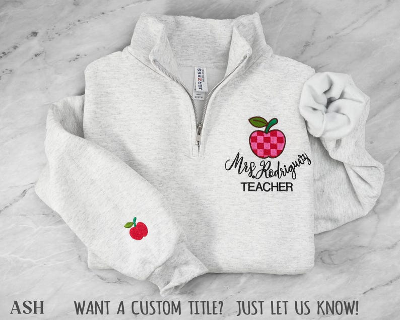 May include: A light gray heather quarter-zip sweatshirt featuring an embroidered red and pink checkered apple design on the chest with the text Mrs. Rodriguez TEACHER below it, and a small red apple embroidered on the right sleeve cuff.