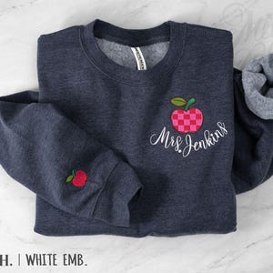 May include: A dark navy heather crewneck sweatshirt featuring an embroidered red and pink checkered apple design on the chest with the white cursive text Mrs. Jenkins underneath. A small red apple is embroidered on the left sleeve cuff.