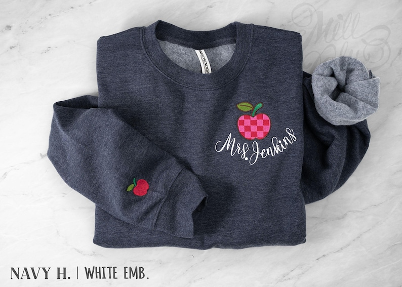 May include: A dark navy heather crewneck sweatshirt featuring an embroidered red and pink checkered apple design on the chest with the white cursive text Mrs. Jenkins underneath. A small red apple is embroidered on the left sleeve cuff.
