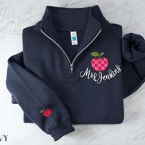 May include: A navy blue quarter-zip sweatshirt featuring a checkered pink and red apple graphic with the embroidered white script text Mrs. Jenkins. A smaller matching apple graphic is embroidered on the left sleeve cuff near the ribbed hem.