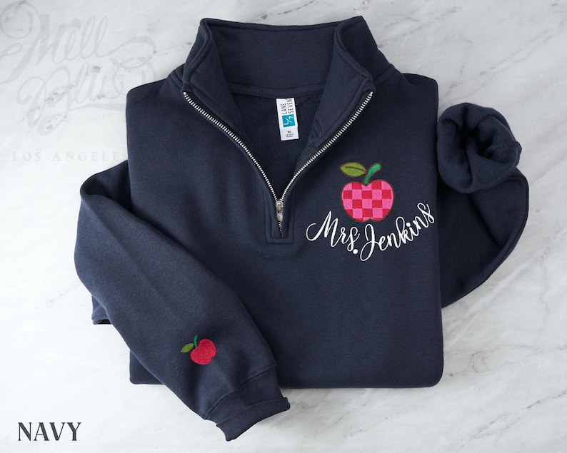 May include: A navy blue quarter-zip sweatshirt featuring a checkered pink and red apple graphic with the embroidered white script text Mrs. Jenkins. A smaller matching apple graphic is embroidered on the left sleeve cuff near the ribbed hem.