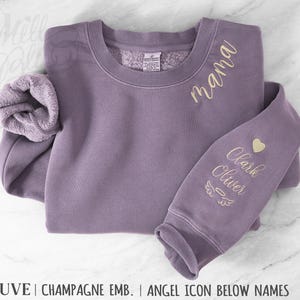 May include: A mauve crewneck sweatshirt featuring gold embroidery. The word mama is embroidered on the upper chest. One sleeve features a heart icon, the names Clark and Oliver, and an angel wing icon with a halo below the names.