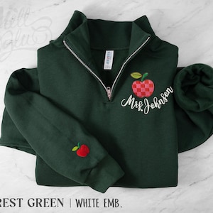May include: A forest green quarter-zip sweatshirt featuring a checkered red apple embroidery on the left chest with the white cursive text Mrs. Johnson below it, and a small red apple embroidered on the right sleeve cuff with a silver zipper.