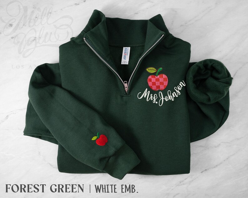 May include: A forest green quarter-zip sweatshirt featuring a checkered red apple embroidery on the left chest with the white cursive text Mrs. Johnson below it, and a small red apple embroidered on the right sleeve cuff with a silver zipper.