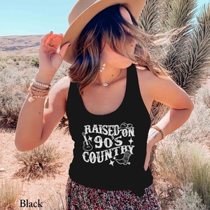 K&ouml;nnte beinhalten: Schwarzes Tanktop mit einem wei&szlig;en Aufdruck, der "Raised on 90's Country" mit einer Gitarre, einem Cowboyhut und Cowboystiefeln zeigt.