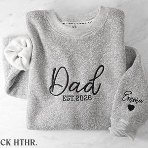 May include: A heather gray crewneck sweatshirt featuring black embroidery on the chest that reads Dad EST. 2026. The left sleeve is embroidered with the name Emma above a small black heart. The interior of the sweatshirt is white.
