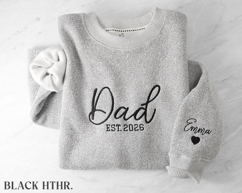 May include: A heather gray crewneck sweatshirt featuring black embroidery on the chest that reads Dad EST. 2026. The left sleeve is embroidered with the name Emma above a small black heart. The interior of the sweatshirt is white.