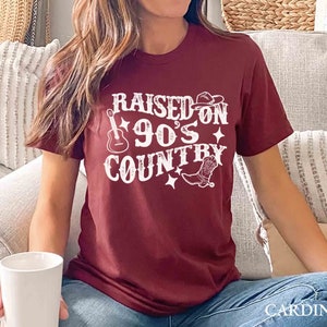 K&ouml;nnte beinhalten: Ein maroonfarbenes T-Shirt mit wei&szlig;em Text, der "Raised on 90's Country" lautet. Der Text ist von einer Gitarre, einem Cowboyhut und einem Cowboystiefel umgeben.