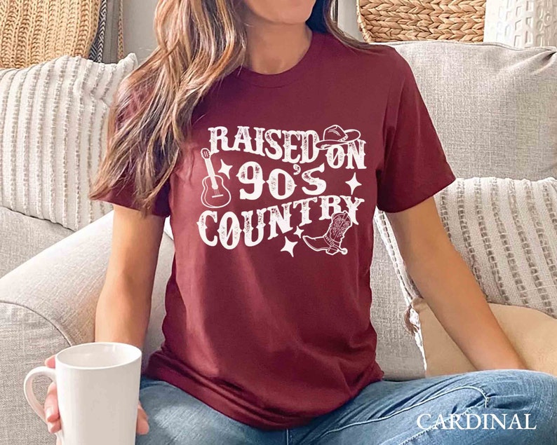 K&ouml;nnte beinhalten: Ein maroonfarbenes T-Shirt mit wei&szlig;em Text, der "Raised on 90's Country" lautet. Der Text ist von einer Gitarre, einem Cowboyhut und einem Cowboystiefel umgeben.