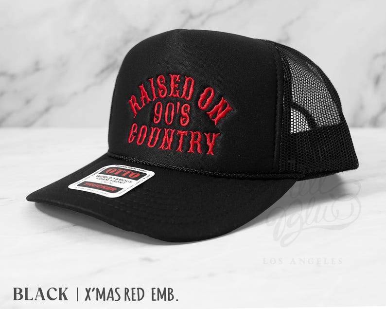 May include: Black trucker hat with a mesh back and a curved brim. The front of the hat features the embroidered text "RAISED ON 90'S COUNTRY" in red. The hat has a snapback closure and a tag that reads "OTTO".