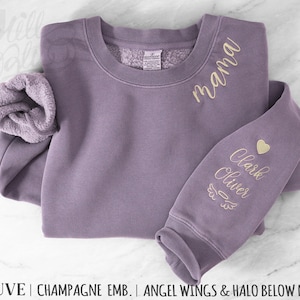 May include: Mauve-colored sweatshirt with the word "mama" embroidered in gold script. The sleeve features the names "Clark Oliver" with a heart, angel wings, and halo design. The sweatshirt is made of a soft material.