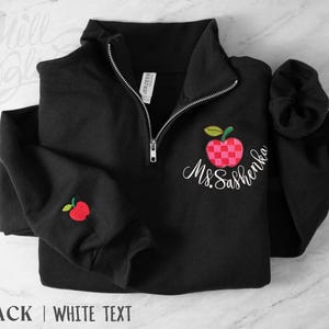 May include: A black quarter-zip sweatshirt featuring an embroidered red and pink checkered apple design on the left chest and a smaller matching apple on the sleeve. The text Ms. Sashenka is embroidered in white script below the chest apple.