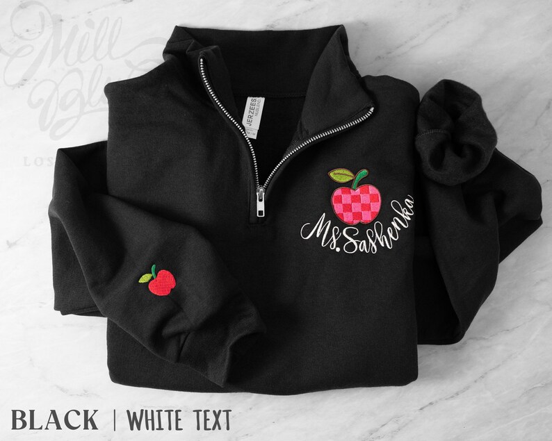 May include: A black quarter-zip sweatshirt featuring an embroidered red and pink checkered apple design on the left chest and a smaller matching apple on the sleeve. The text Ms. Sashenka is embroidered in white script below the chest apple.