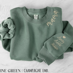 May include: Alpine green sweatshirt with champagne embroidery. The word "mama" is embroidered on the chest, and names "Avery, Addie, Robbie" are embroidered on the sleeve, with a heart symbol. A matching scrunchie is included.