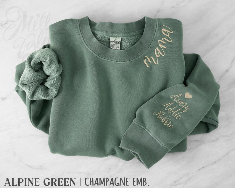May include: Alpine green sweatshirt with champagne embroidery. The word "mama" is embroidered on the chest, and names "Avery, Addie, Robbie" are embroidered on the sleeve, with a heart symbol. A matching scrunchie is included.