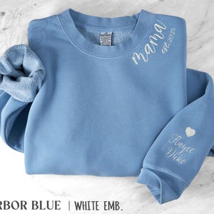 May include: A light blue sweatshirt with white embroidered text reading "mama est.2025" on the upper chest. The sleeve features a heart and the name "Hayce Duke". The sweatshirt is labeled "Harbor Blue | White Emb."