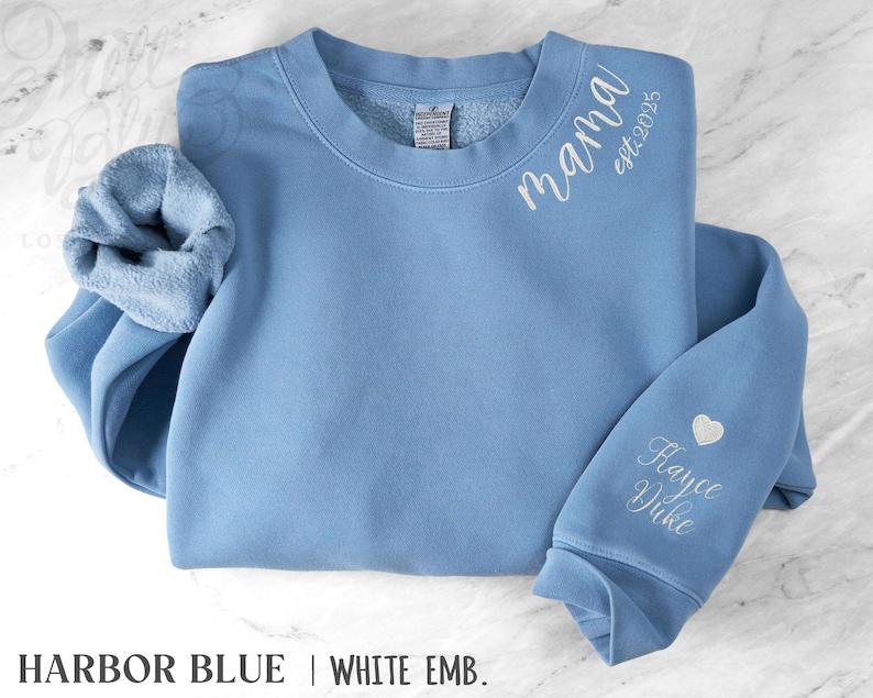 May include: A light blue sweatshirt with white embroidered text reading "mama est.2025" on the upper chest. The sleeve features a heart and the name "Hayce Duke". The sweatshirt is labeled "Harbor Blue | White Emb."