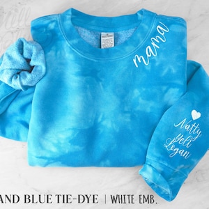 May include: A turquoise tie-dye sweatshirt with white embroidered text reading "mama" on the chest and names on the sleeve. A white heart is embroidered above the names. A matching scrunchie is included. The sweatshirt is a crew neck.