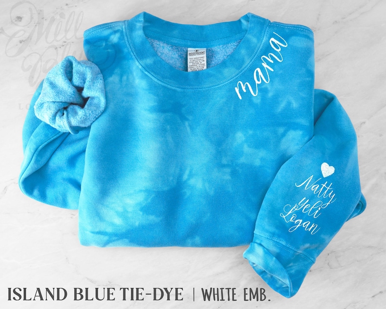 May include: A turquoise tie-dye sweatshirt with white embroidered text reading "mama" on the chest and names on the sleeve. A white heart is embroidered above the names. A matching scrunchie is included. The sweatshirt is a crew neck.