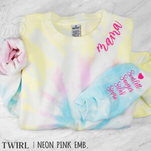 May include: A pastel tie-dye sweatshirt in yellow, pink, and blue. The word "mama" is embroidered in neon pink. The sleeve cuff is light blue with embroidered names and a heart. The text "BOHO TWIRL | NEON PINK EMB." is at the bottom.