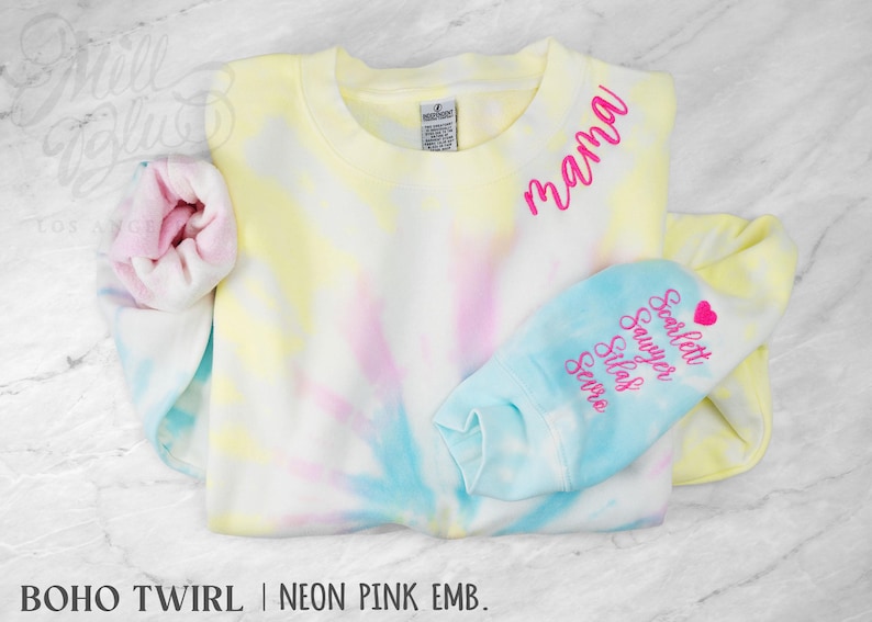 May include: A pastel tie-dye sweatshirt in yellow, pink, and blue. The word "mama" is embroidered in neon pink. The sleeve cuff is light blue with embroidered names and a heart. The text "BOHO TWIRL | NEON PINK EMB." is at the bottom.