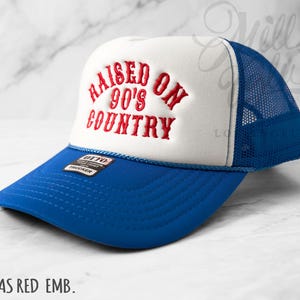 May include: A blue and white trucker hat with the embroidered text "RAISED ON 90'S COUNTRY" in red. The hat features a blue brim and mesh back, with a white front panel. The hat has a small tag that says "OTTO TRUCKER".