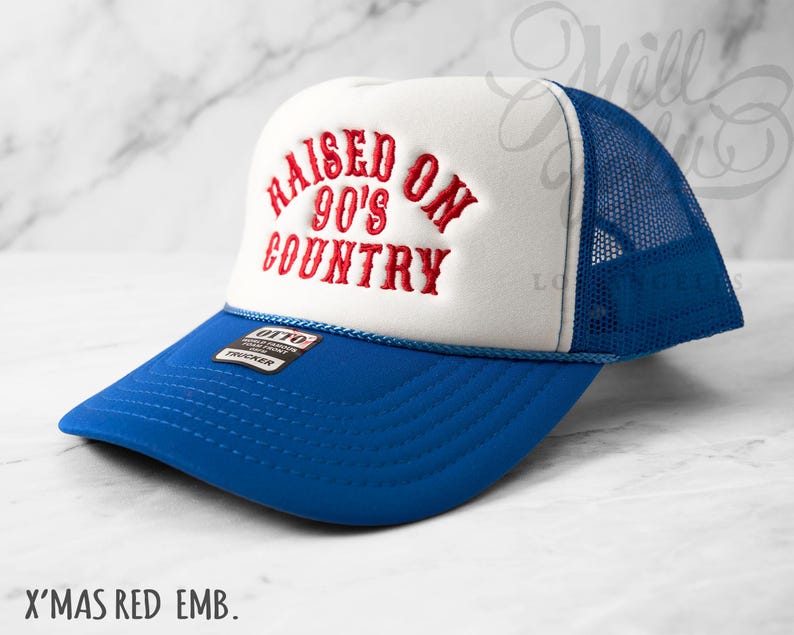 May include: A blue and white trucker hat with the embroidered text "RAISED ON 90'S COUNTRY" in red. The hat features a blue brim and mesh back, with a white front panel. The hat has a small tag that says "OTTO TRUCKER".