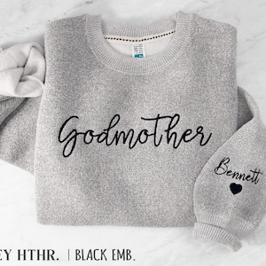May include: A heather grey crewneck sweatshirt featuring the word Godmother embroidered in black script across the chest. The right sleeve displays the name Bennett in black script above a small black embroidered heart icon.