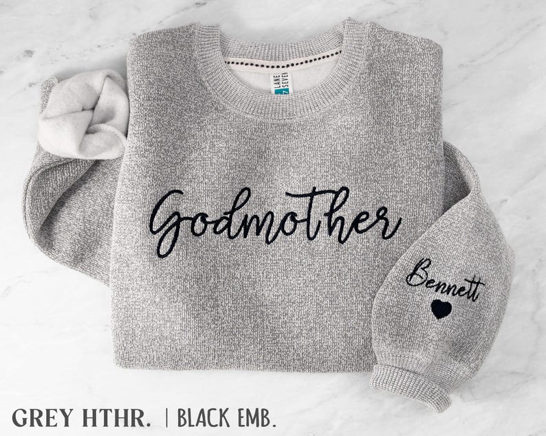 May include: A heather grey crewneck sweatshirt featuring the word Godmother embroidered in black script across the chest. The right sleeve displays the name Bennett in black script above a small black embroidered heart icon.
