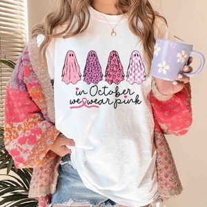 May include: White t-shirt with a pink ribbon and the text "in October we wear pink". Four pink ghosts are printed on the shirt, each with a different pattern: polka dots, leopard print, hearts, and checkered.