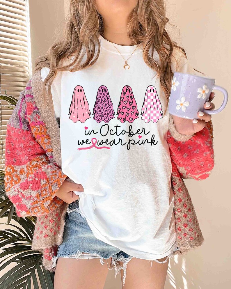 May include: White t-shirt with a pink ribbon and the text "in October we wear pink". Four pink ghosts are printed on the shirt, each with a different pattern: polka dots, leopard print, hearts, and checkered.