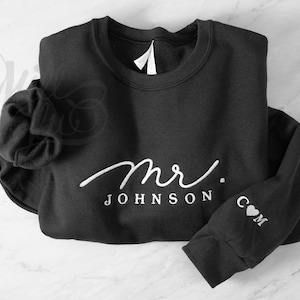 May include: A black long-sleeve sweatshirt featuring white embroidered text that reads mr. JOHNSON on the chest. The sleeve includes the embroidered letters C and M separated by a small heart symbol. The garment has a crew neckline.