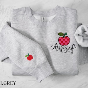 May include: A light heather gray crewneck sweatshirt featuring an embroidered red and pink checkered apple with a green leaf on the chest, accompanied by the embroidered black script text Mrs. Boyer. A smaller matching apple is on the sleeve.