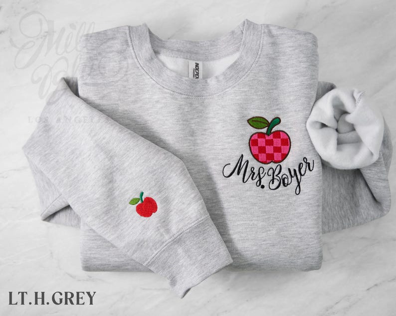 May include: A light heather gray crewneck sweatshirt featuring an embroidered red and pink checkered apple with a green leaf on the chest, accompanied by the embroidered black script text Mrs. Boyer. A smaller matching apple is on the sleeve.