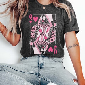 K&ouml;nnte beinhalten: Ein dunkelgraues Heather-T-Shirt mit einer pinkfarbenen und schwarzen Grafik der Herzdame aus Spielkarten. Die Dame tr&auml;gt eine Sonnenbrille und h&auml;lt ein Glas Wein in der Hand. Der Text "More Oceans" steht auf dem rechten &Auml;rmel des Shirts.