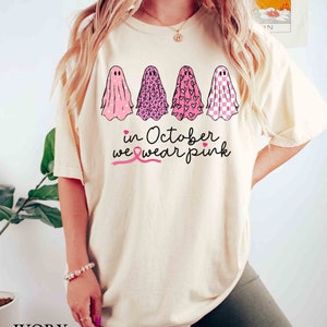 May include: A cream-colored t-shirt with a pink ribbon, four ghosts with pink, leopard, heart, and checkered patterns, and the text "in October we wear pink".