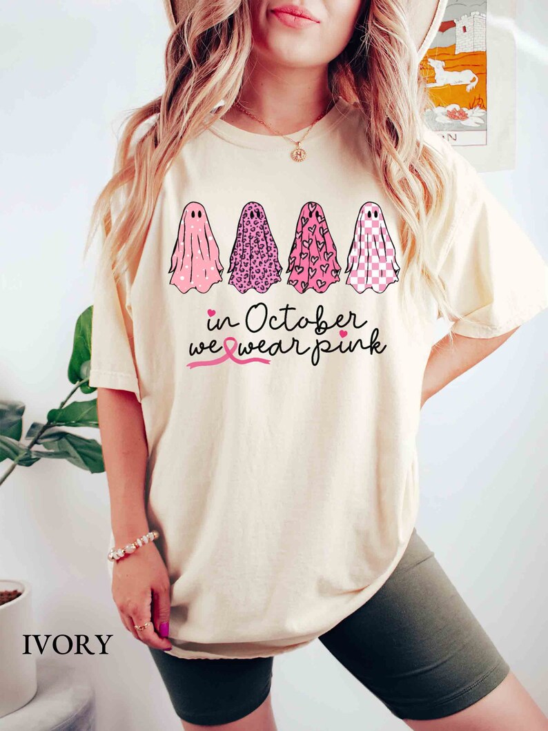 May include: A cream-colored t-shirt with a pink ribbon, four ghosts with pink, leopard, heart, and checkered patterns, and the text "in October we wear pink".