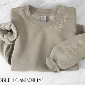May include: A beige sweatshirt with champagne-coloured embroidery. The word "mama" is embroidered on the chest, and a name and heart are embroidered on the sleeve. The sweatshirt has a crew neck and ribbed cuffs.