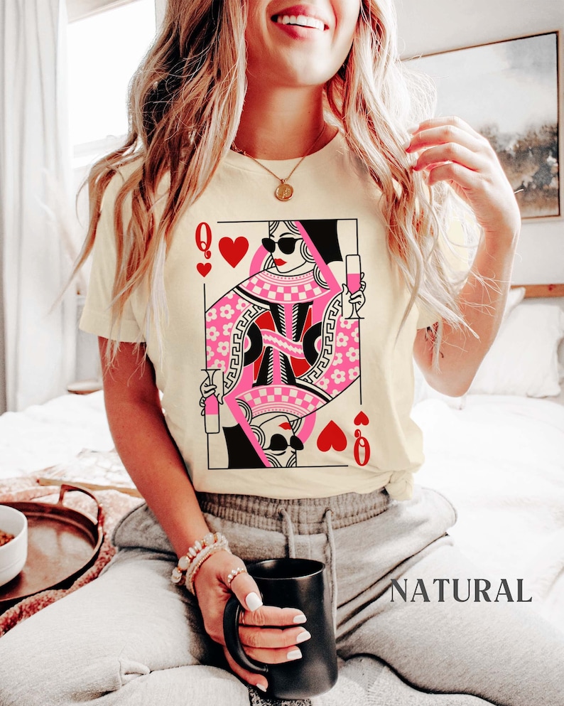 K&ouml;nnte beinhalten: Ein wei&szlig;es T-Shirt mit einem Aufdruck der Herzdame aus einem Kartenspiel. Die Dame tr&auml;gt eine Sonnenbrille und h&auml;lt ein Glas Champagner in der Hand. Der Text "NATURAL" ist in der rechten unteren Ecke des Bildes zu sehen.
