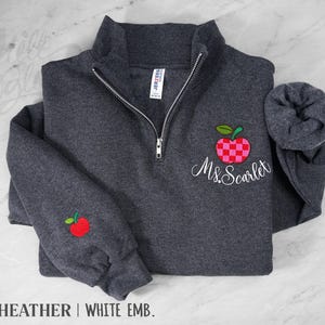 May include: A dark heather gray quarter-zip sweatshirt featuring a silver zipper and a white embroidered name Ms. Scarlet. An apple with a red and pink checkered pattern is embroidered above the name, and a small red apple is on the sleeve.