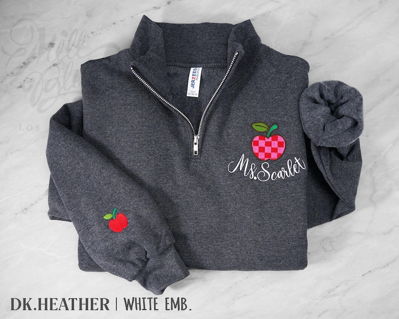 May include: A dark heather gray quarter-zip sweatshirt featuring a silver zipper and a white embroidered name Ms. Scarlet. An apple with a red and pink checkered pattern is embroidered above the name, and a small red apple is on the sleeve.