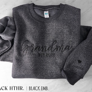 May include: A dark charcoal heather gray crewneck sweatshirt featuring black embroidery on the chest that reads Grandma EST. 2026. The sleeve includes an embroidered black heart and a name. The interior is lined with a soft fleece material.