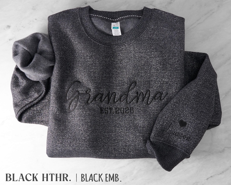 May include: A dark charcoal heather gray crewneck sweatshirt featuring black embroidery on the chest that reads Grandma EST. 2026. The sleeve includes an embroidered black heart and a name. The interior is lined with a soft fleece material.